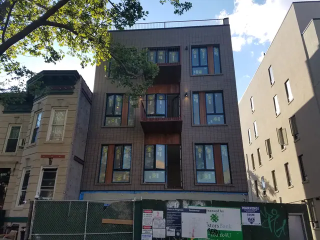Street view of 1503 Jefferson Avenue during visible construction, showing a dark brick facade, central projecting balconies, window labels, and a green perimeter fence.