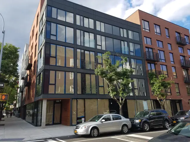 Street photograph of 152 Manhattan Avenue showing a dark-framed glazed corner, red-brick elevations and recessed ground-floor entrance.