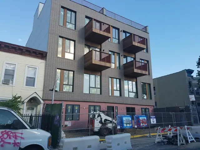 Angled street view of 1534 Putnam Avenue, with a gray-brick facade, four projecting balconies, a pink ground-floor finish, and construction barriers.