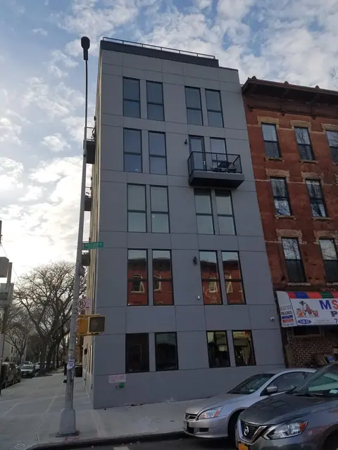 Street photograph of 182 Malcolm X Boulevard showing the narrow gray-clad facade, a projecting balcony, rooftop railing and neighboring brick building.