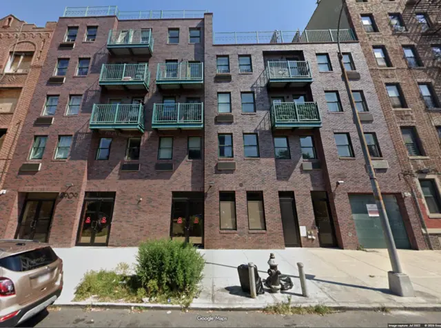 Street View photograph of a broad brick building with green-painted balconies, rooftop railings and a stepped roofline above ground-floor entrances.