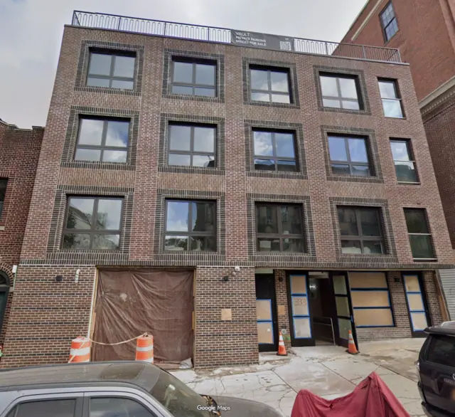 Street View photograph of a four-story brick building with large dark-framed windows, a rooftop railing and a partially screened ground-floor opening.