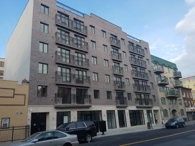 Three-quarter street view of 667 Myrtle Avenue, showing a broad brick facade, repeated black-railed balconies, glazed ground-floor openings, and neighboring buildings.