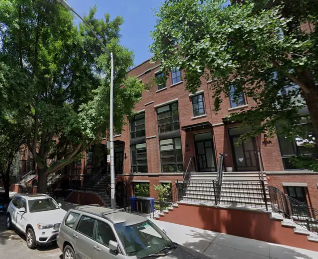 Street View photograph of red-brick townhouses with raised front stoops, black railings and tall dark window bays beneath mature street trees.